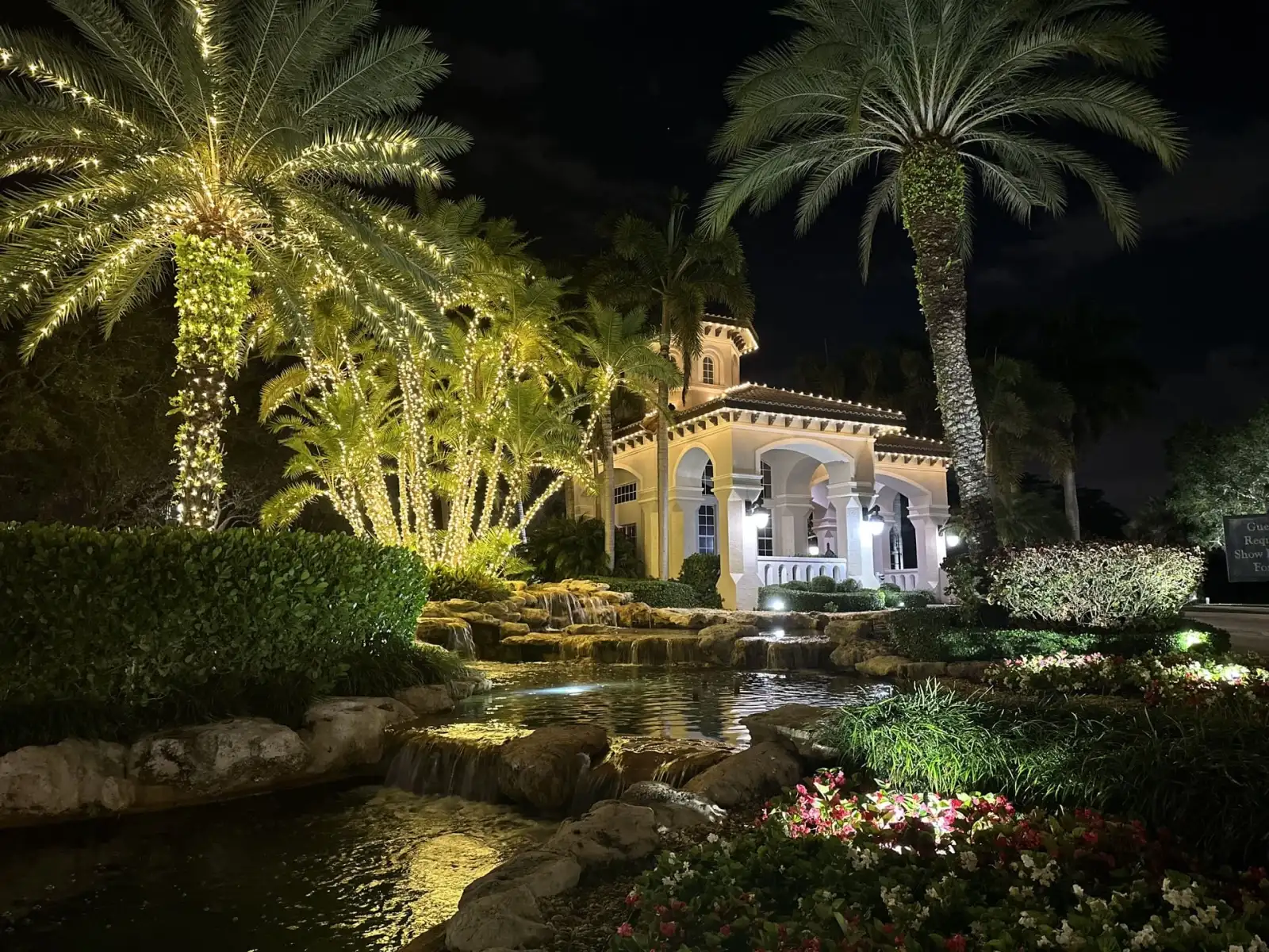 Mediterranean clubhouse with palm trees wrapped in warm white holiday lights beside a lit waterfall at night